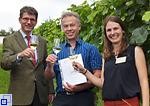 Dr. Martin Barth, der Preisträger Daniel Feuerstein und Andrea Gierden stehen vor Weinreben und stoßen mit dem ausgezeichneten Sekt an. 